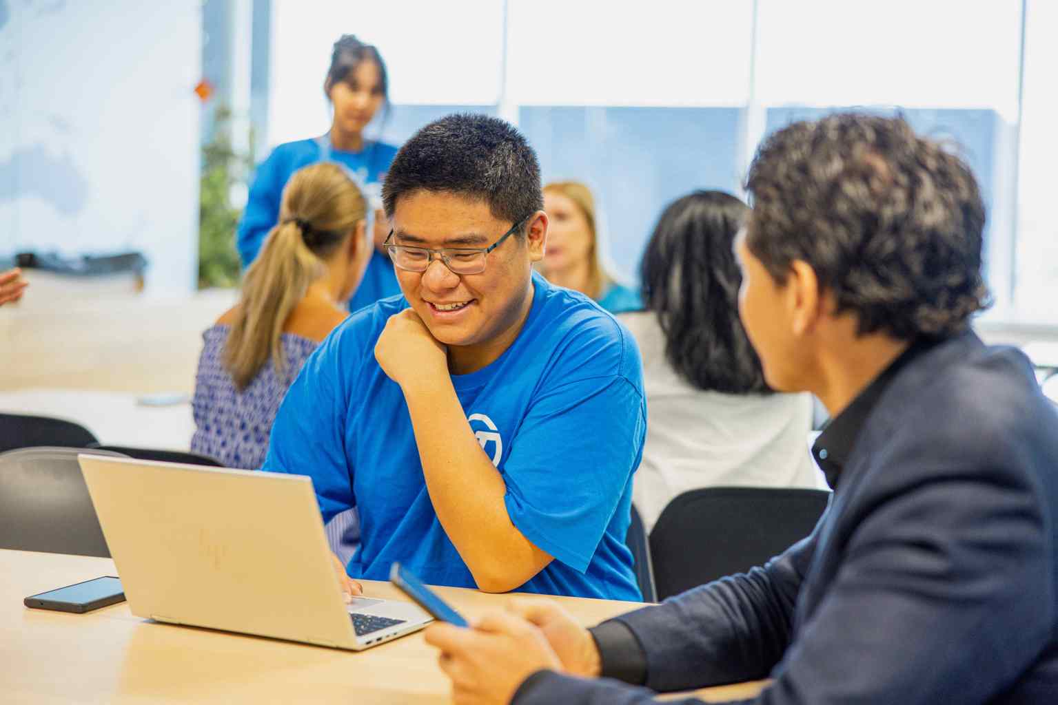 A man in a blue shirt sits at a laptop looking down at the screen. A man whose face is not shown sits watching him, holding his mobile phone.